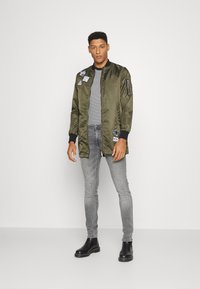 Chaqueta bomber en verde oliva con ribetes negros, con parches, bolsillos con cremallera, combinada con una camiseta de rayas, jeans skinny grises y botas negras.