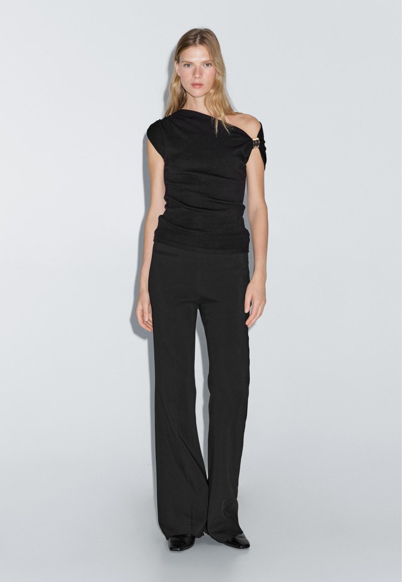 Svart off-shoulder topp med draperat tyg och en guldig accent. Kombinerad med högmidjade, vida svarta byxor, som båda har en jämn textur.