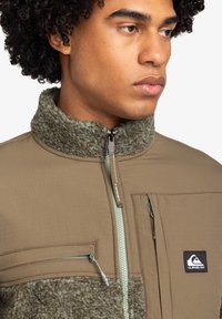 Fleece- och nylonjacka i beige och grön, med dragkedja framtill, hög krage och bröstficka med logodetalj. Texturerade detaljer finns.