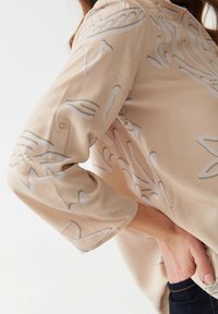 Bluse aus hellbeigem Stoff mit abstrakten grauen und weißen Mustern. Dreiviertelärmel mit Knopfdetail am Bündchen.