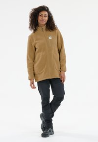 Beige Fleecejacke mit hohem Kragen, Fronttaschen und einem kleinen Logo-Patch. Kombiniert mit dunklen, schmal geschnittenen Outdoor-Hosen und robusten schwarzen Stiefeln.