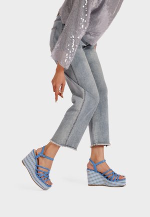 Persona che indossa sandali con zeppa a cinturini azzurri chiari, jeans grigi corti e un maglione grigio con decorazioni di paillettes sulle maniche.