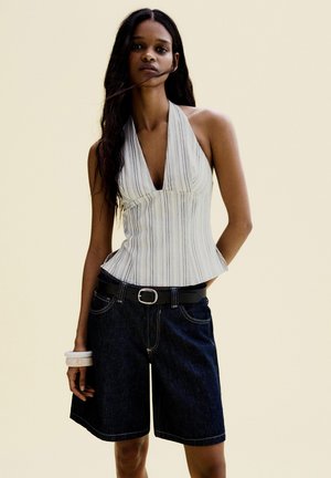 Nő ujjatlan csíkos pántos felsőt, sötét kék farmer térdig érő rövidnadrágot fekete övvel, valamint fehér karpereceket visel a bal csuklóján.