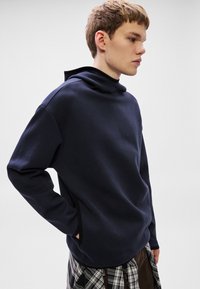 Námořnicky modrá mikina s vysokým límcem, bočními kapsami a texturovanou látkou. Má volný střih a minimalistický design.