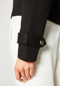 Czarna, teksturowana rękaw kurtki z mankietem z guzikiem, w zestawieniu z białymi spodniami z tkaniny.