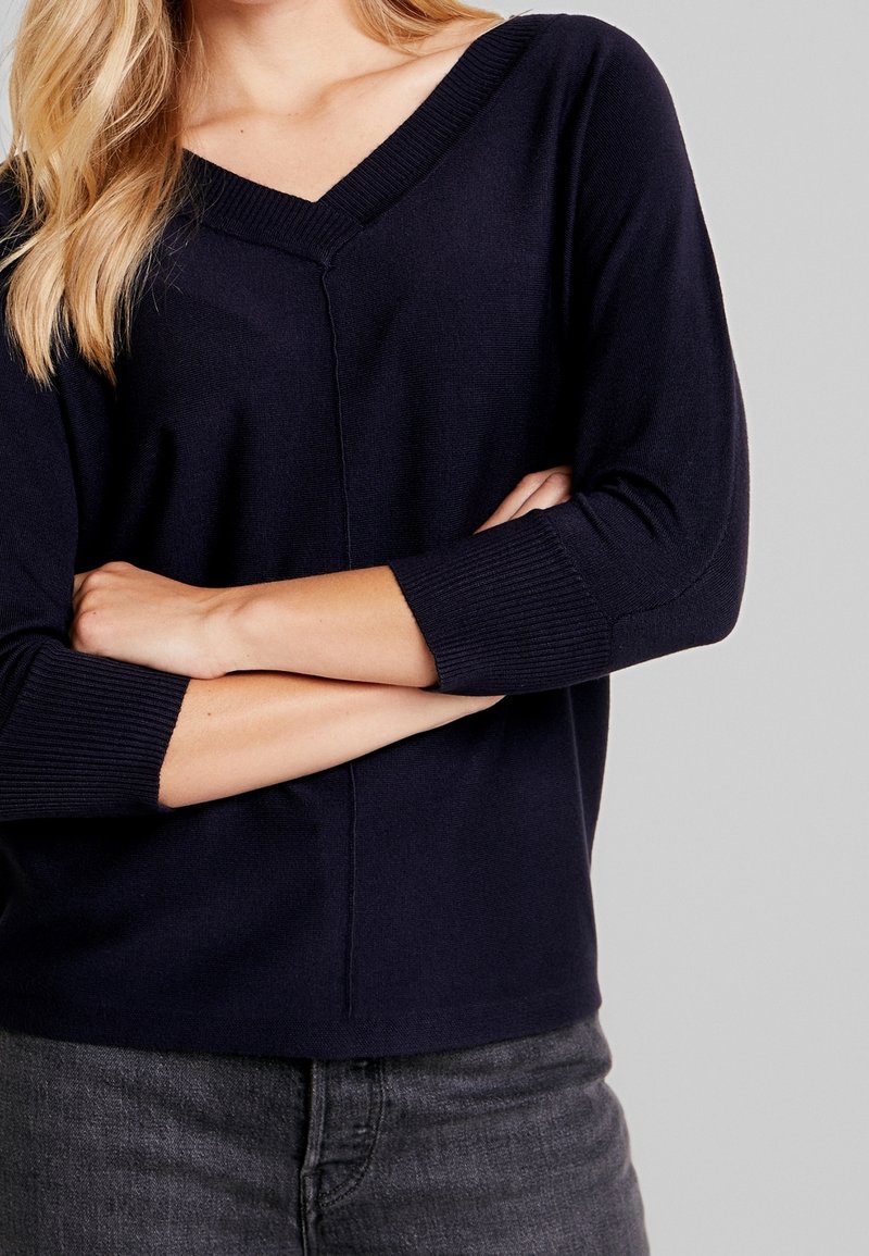 Femme aux bras croisés portant un pull noir à col en V et un jean gris sur un fond gris uni.