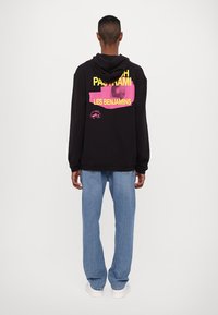 Black hoodie with a large graphic print on the back in pink and yellow text. Loose fit, features a hood, paired with light blue jeans.