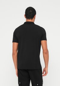 Muž vidieť zozadu, oblečený v čiernom polokošeli s krátkym rukávom a čiernych nohaviciach s vreckami, na bielom neutrálnom pozadí.