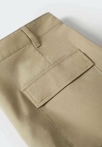 Pantalon beige avec passants de ceinture et une grande poche à rabat rectangulaire avec des coutures visibles sur le côté.