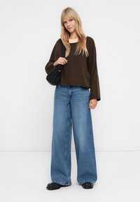 Suéter castanho oversized, jeans de perna larga azuis, bolsa preta e sapatos pretos robustos. Texturas suaves, ajuste descontraído e silhueta relaxada.