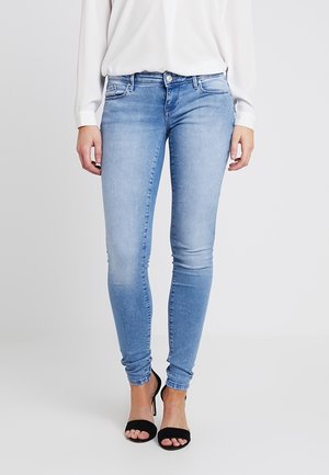 Mujer con vaqueros ajustados azul claro, blusa blanca de manga larga y sandalias de tacón negro con los dedos al aire, de pie frente a un fondo liso.