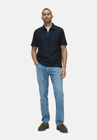 Marineblauwe T-shirt met korte mouwen en een ritssluiting bij de nek, gecombineerd met lichtblauwe rechte jeans en donkerbruine schoenen.