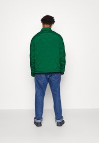 Giacca a vento verde imbottita con motivo a rombi, abbinata a jeans blu e scarpe nere. La giacca ha un colletto ed è di lunghezza media.