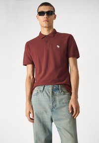 Weinrotes Poloshirt mit einer Drei-Knöpfe-Leiste und einem kleinen weißen Logo. Kombiniert mit hellblauen Jeans, die eine lässige Passform zeigen.