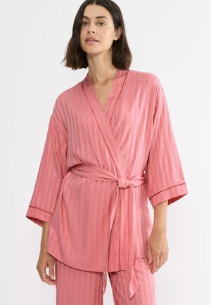 Frau mit schulterlangem dunklem Haar, die einen rosa gestreiften Morgenmantel mit Gürtel und passende Hosen vor einem weißen Hintergrund trägt.