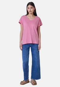 Růžové tričko s krátkým rukávem a výstřihem do V vyrobené z lehké tkaniny, spárované s modrými kalhotami s širokými nohavicemi a béžovými sandály s dekorativními akcenty.