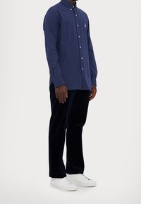 Mörkblå skjorta med knappar och vita prickar, ihopparad med mörka sammetsbyxor och vita sneakers. Har en krage och diskret logotypdetalj.