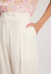 Pantaloni a gamba larga bianchi con un design sartoriale, caratterizzati da pieghe frontali e tasche laterali. Tessuto morbido e chiusura con un solo bottone in vita.