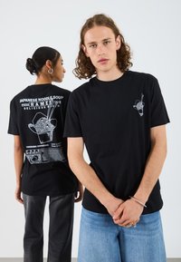 Svart t-skjorter med grafiske design: foran har små print av spisepinner og nudler; baksiden viser en større illustrasjon av en ramenboks med tekst.