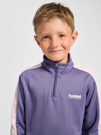 Jonge jongen met kort lichtbruin haar, gekleed in een paarse sportjas met een kwart ritssluiting en het logo "hummel performance", licht glimlachend.