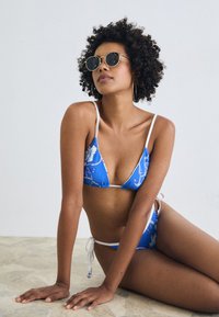 Blauer Blumenbikini mit weißen Trägern, bestehend aus einem Dreieckstop und Seitenschnüren. Model trägt runde Sonnenbrillen mit einem goldenen Rahmen.