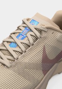 Μπεζ πλεκτό παπούτσι για τρέξιμο Nike με κρέμ κορδόνια και μπλε ετικέτα που γράφει "Terra Kiger", με μοβ λογότυπο Nike στο πλευρό.