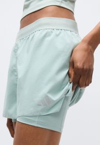 Shorts de sport vert menthe clair avec poche cachée et ceinture élastique. Comprend trois rayures grises sur le côté inférieur gauche. Texture lisse.