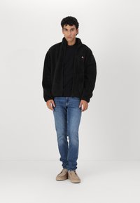 Chaqueta de forro polar negra con cierre de cremallera, cuello alto y parche de logo, combinada con jeans azules y zapatos beigs. Tela suave con una textura blandita.