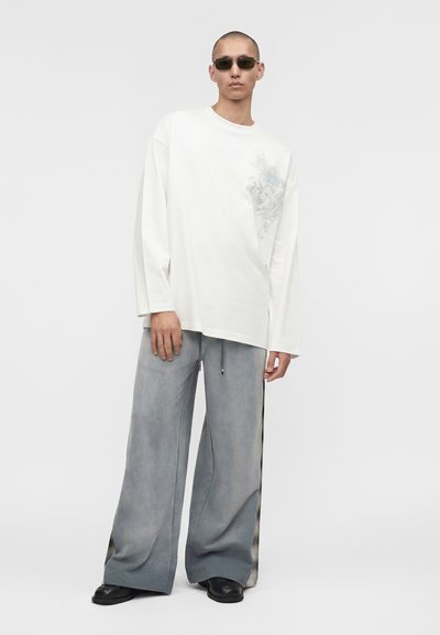 Άτομο που φοράει υπερμεγέθη λευκό πουκάμισο με μακριά μανίκια και διακριτικό λουλουδάτο σχέδιο, φαρδιά γκρι παντελόνια με πλαϊνές ρίγες, μαύρα παπούτσια και σκούρα γυαλιά ηλίου.