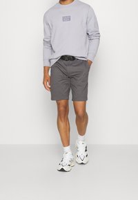 Ljuslila sweatshirt med en logotyp, grå shorts och vita sportskor. Outfitten har en avslappnad passform och minimalistiska designelement.