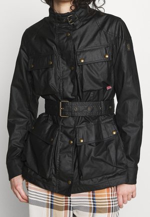 Persona que lleva una chaqueta negra con cinturón, múltiples botones de latón y un pequeño parche de la Union Jack en el pecho, combinada con pantalones a cuadros.