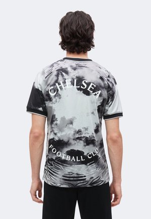 Mann trägt ein schwarz-weißes Tie-Dye Chelsea Football Club Trikot, von hinten mit beiden Armen entspannt nach unten gezeigt.