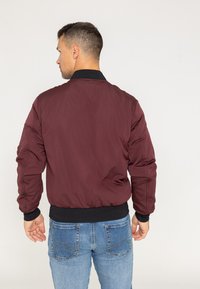 Burgundowa kurtka bomber z czarnymi ściągaczami na mankietach i u dołu; gładka struktura; ocieplana konstrukcja; pokazana z tyłu na tle niebieskich dżinsów.