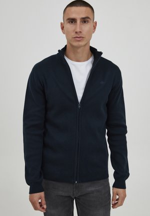 Maglioncino zip navy, materiale lavorato a maglia, design aderente, polsini e orlo a coste, con un piccolo logo sul petto. Indossato sopra una camicia bianca.