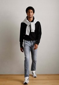Zwarte lange mouwen shirt, grijze sweatshirt over de schouders gedrapeerd, lichtblauwe jeans en wit-zilveren sneakers, staand op een houten vloer.
