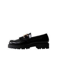Mocasines de charol negro con un detalle de hebilla dorada y una suela de goma gruesa. Presentan un acento de flecos en la parte delantera y un diseño elegante.