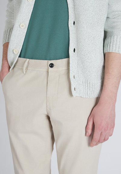 Lichtgrijze gebreide cardigan over een teal shirt, gecombineerd met beige broeken met een knoopsluiting, zijzakken en een gladde textuur.