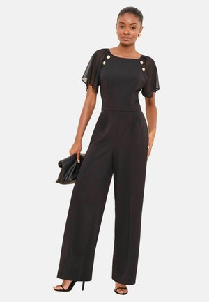 Schwarzer Jumpsuit mit transparenten kurzen Ärmeln, quadratischem Ausschnitt und goldenen Knöpfen. Weit geschnittenes Design mit glatter Textur, ergänzt durch eine schwarze Clutch.