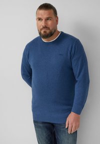 Blauer Rundhals-Pullover aus einem weichen, strukturierten Strick. Mit gerippten Bündchen und Saum. Kombiniert mit Jeans. Dezentes Logo auf der Brust.