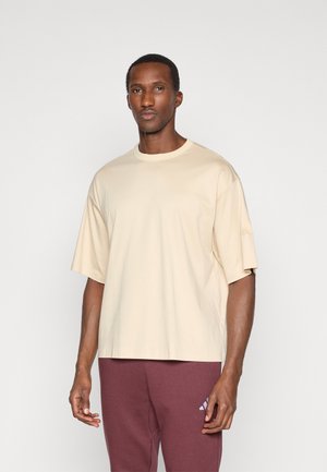 Beige übergroßes T-Shirt aus weichem Stoff, mit rundem Ausschnitt und kurzen Ärmeln. Kombiniert mit bordeauxroten Jogginghosen, die ein Logo zeigen.