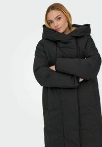 Černá péřová bunda s velkou kapucí, s prošívaným designem, dvěma bočními kapsami a uvolněným, oversized střihem. Hladká textura.