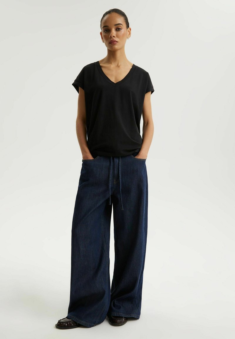 Kobieta stojąca z rękami w kieszeniach, ubrana w czarną bluzkę z krótkim rękawem i dekoltem w serek, ciemne szerokie jeansy oraz czarne buty na białym tle.