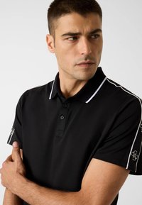 Czarna koszulka polo z trzema guzikami, białym kołnierzykiem i paskami po bokach oraz akcentami logo na rękawach. Wykonana z teksturowanej tkaniny.