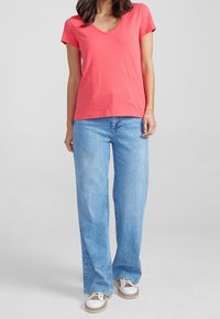 T-shirt a v in corallo realizzata in tessuto morbido, abbinata a jeans a gamba larga di un blu chiaro e sneakers bianche con accenti marroni. Design semplice, senza fantasie.
