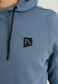 Světle modrá mikina s kapucí vyrobená z texturované tkaniny, obsahující černé šňůrky a malou černou nášivku s bílým geometrickým vzorem.