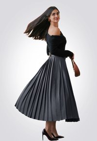 Top nero off-shoulder abbinato a una gonna plissettata grigia, evidenziando un design fluido e una texture liscia. Inclusi tacchi alti neri e una borsa marrone.
