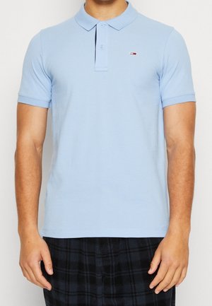 Homme portant un polo à manches courtes bleu clair avec un petit logo rouge et blanc sur la poitrine et un pantalon à carreaux foncé.