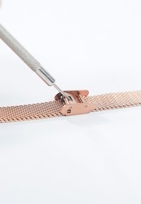 Ajustement d'une boucle de bracelet de montre en maille métallique en or rose à l'aide d'un outil de précision sur une surface blanche.