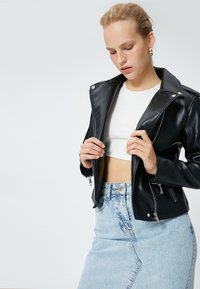 Svart konstläder bikerjacka med silverdetaljer, fickor med dragkedja och klippt krage, stylad över en vit croptop och ljus denimkjol.