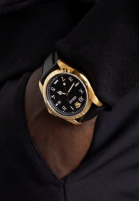 Arany színű óra fekete számlappal, fekete bőrszíjjal és fehér óraincsekkel. Domború lünetta és logó látható az alján.